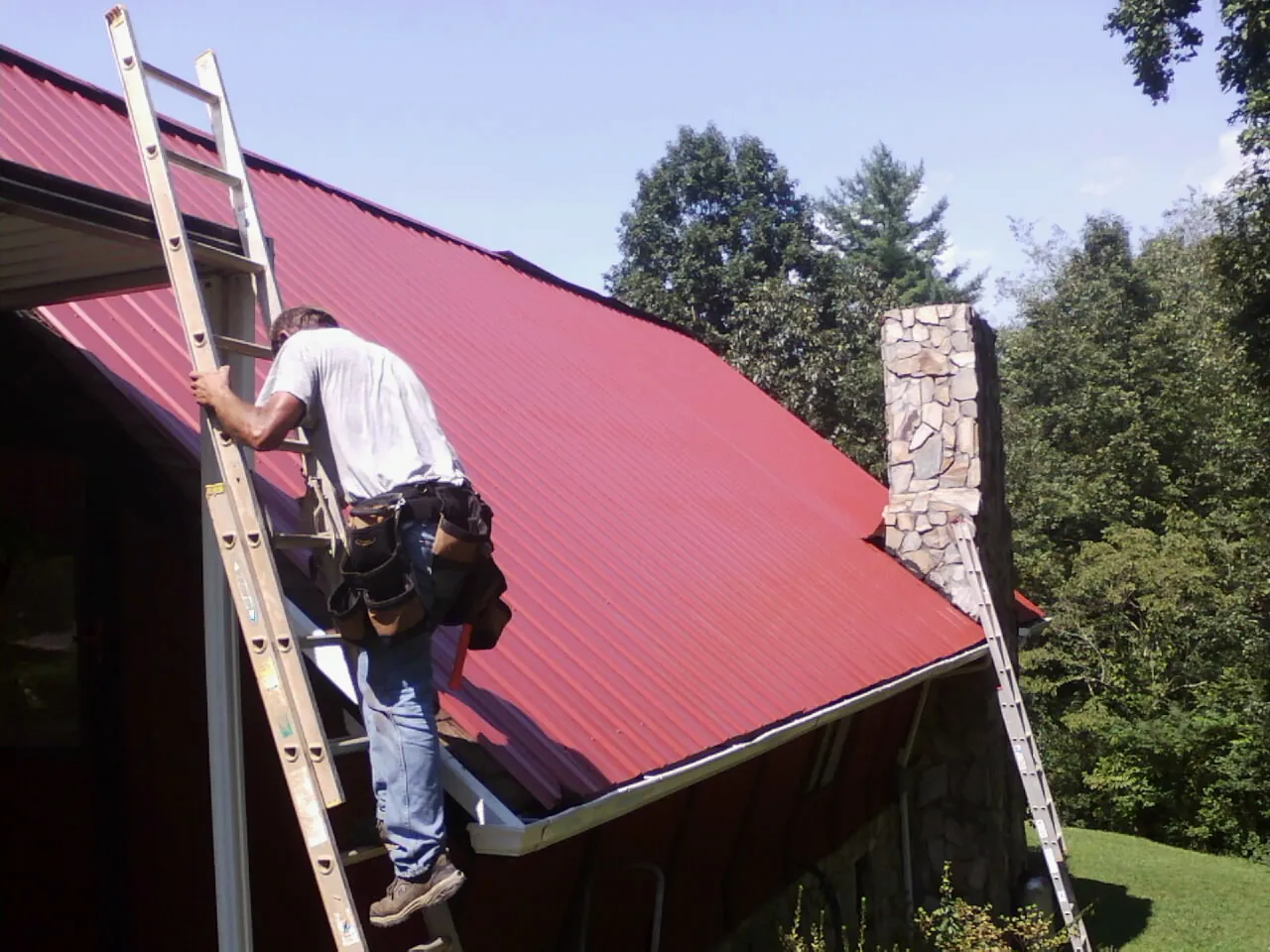 Roofer on a ladder beside a red metal roof and stone chimney — Soffit & Fascia Repair by Roofing Brothers in Lockbourne